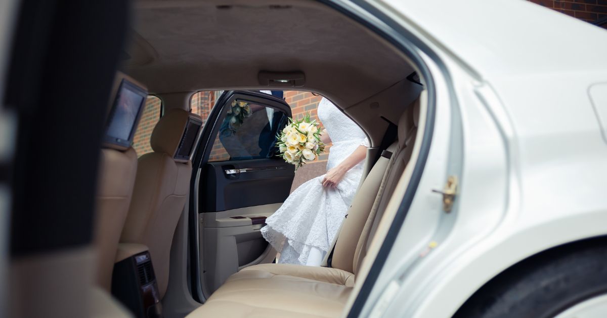 Bride in a white wedding dress holding a bouquet as she steps out of the back seat of a luxury car with a beige leather interior.