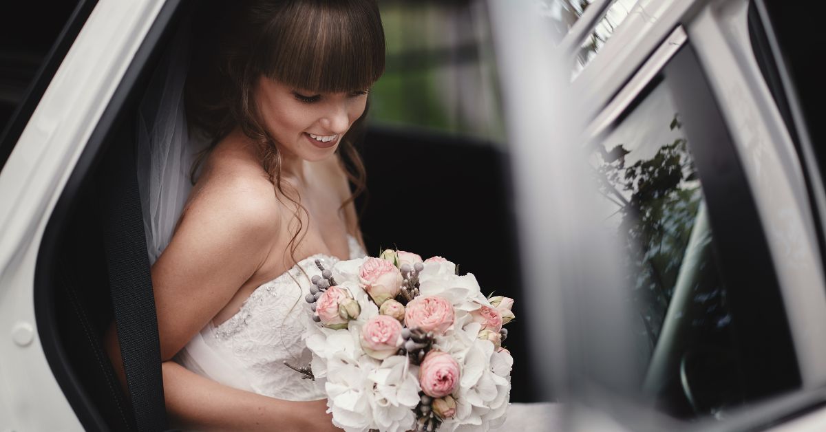 Smiling bride in a white wedding dress sitting inside a car and holding a bouquet of white and pink flowers as she prepares to step out.