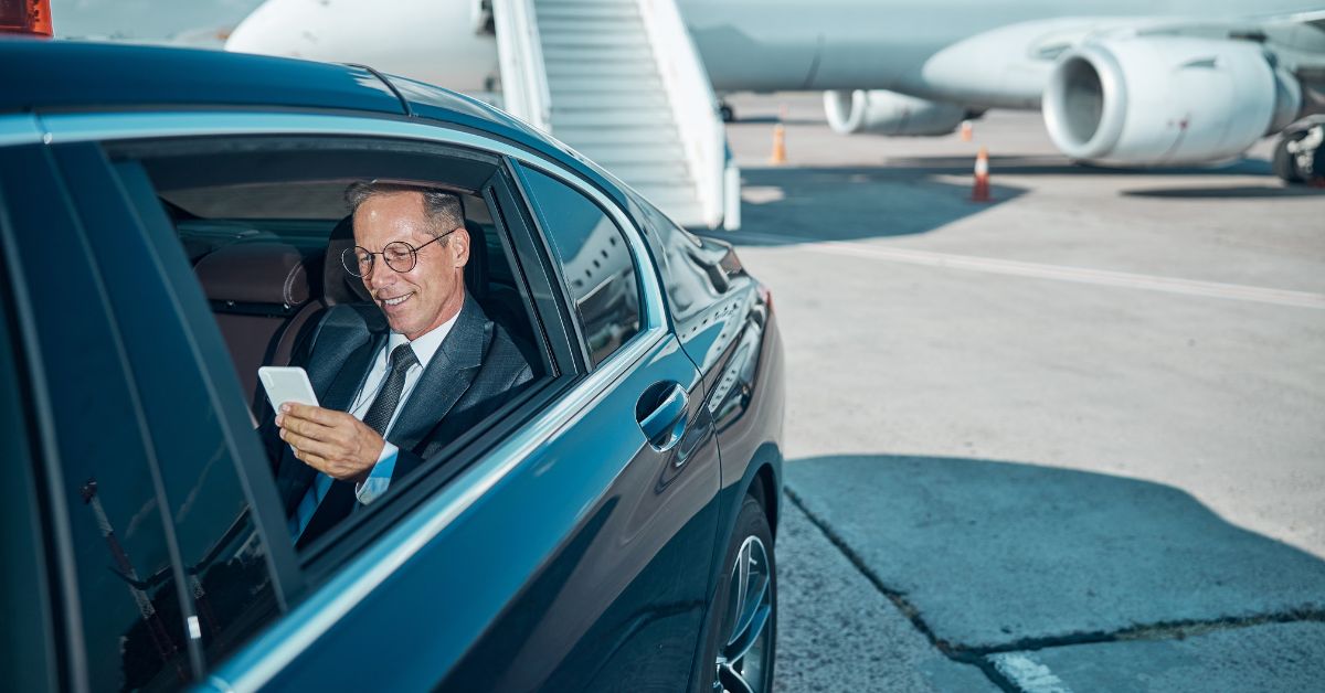 Businessman in a suit sitting in the back seat of a car using a smartphone on an airport runway, with a private jet and boarding stairs visible in the background.