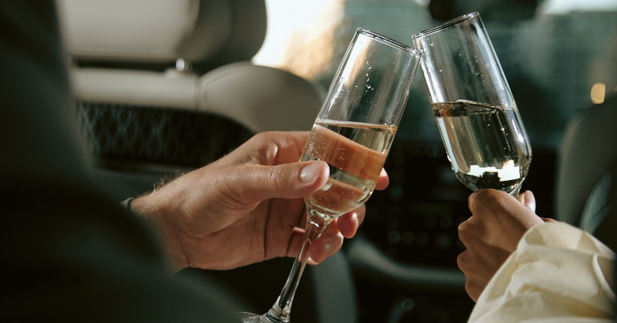 Close-up of two people clinking champagne flutes filled with sparkling wine while sitting in the back seat of a car, celebrating during a ride.