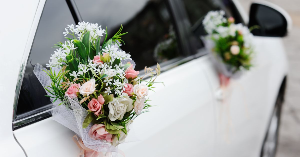 Close-up of a white wedding car decorated with a bouquet of pink and white flowers attached to the door handle, with another matching floral arrangement visible on the rear door.
