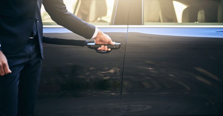 A person in a dark formal suit opens the door of a sleek black luxury car, suggesting a chauffeur service or professional transportation.