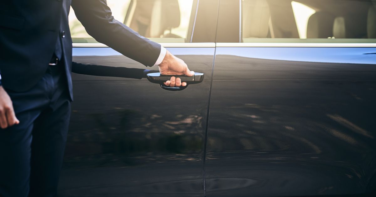 A person in a dark formal suit opens the door of a sleek black luxury car, suggesting a chauffeur service or professional transportation.