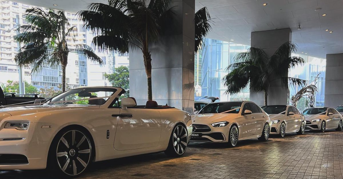 A row of luxury white cars parked beneath a modern covered drive, including a Rolls-Royce convertible in the foreground and several Mercedes sedans behind it, with tall palm trees and city buildings visible in the background.