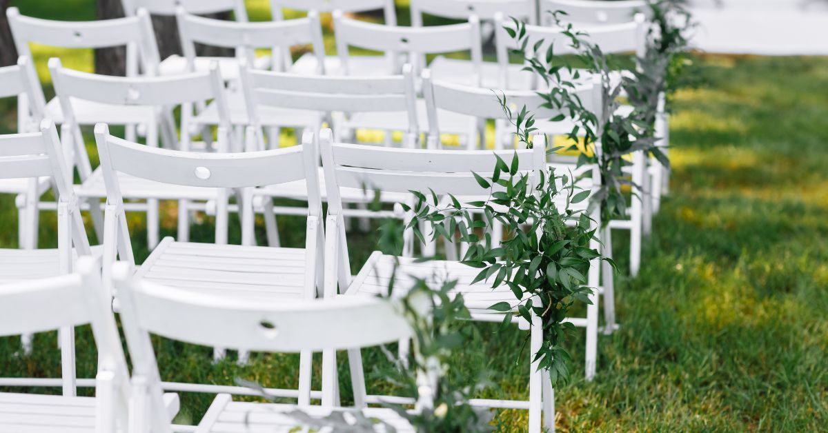 Rows of white folding chairs arranged outdoors on green grass, decorated with sprigs of greenery along the aisle, suggesting a setup for a wedding or event.